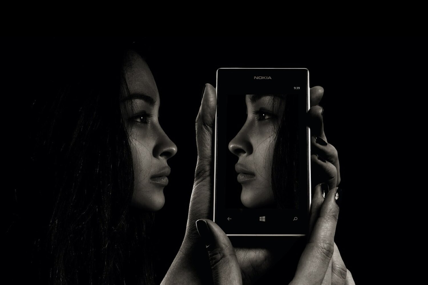 KL Tarot. A person holds a smartphone, which displays a close-up profile of their face on the screen, mirroring their actual profile visible next to it, all set against a dark background.