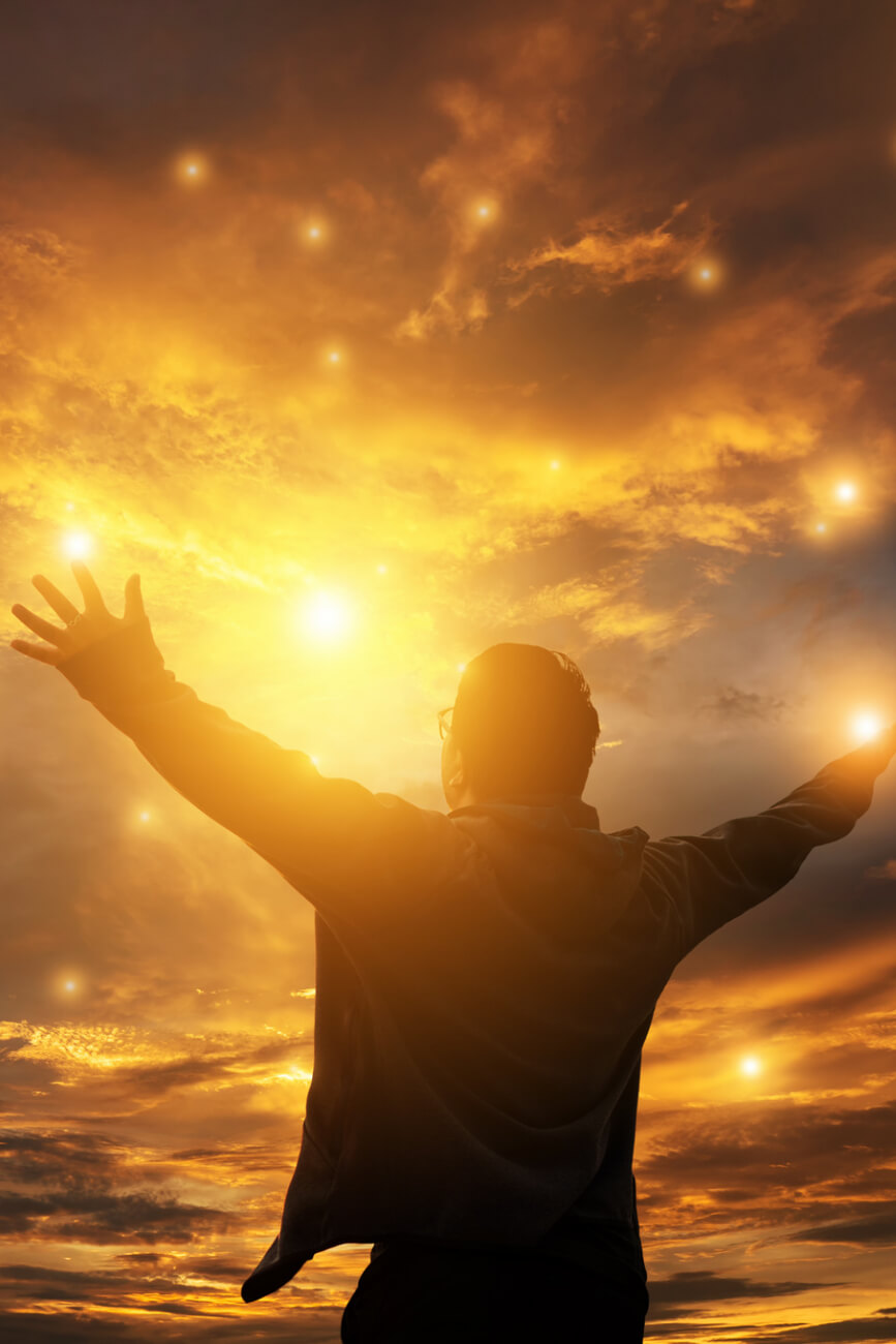 KL Tarot. Silhouette of a person with arms raised, standing against a dramatic sky with vibrant orange and yellow clouds, illuminated by multiple bright light spots.