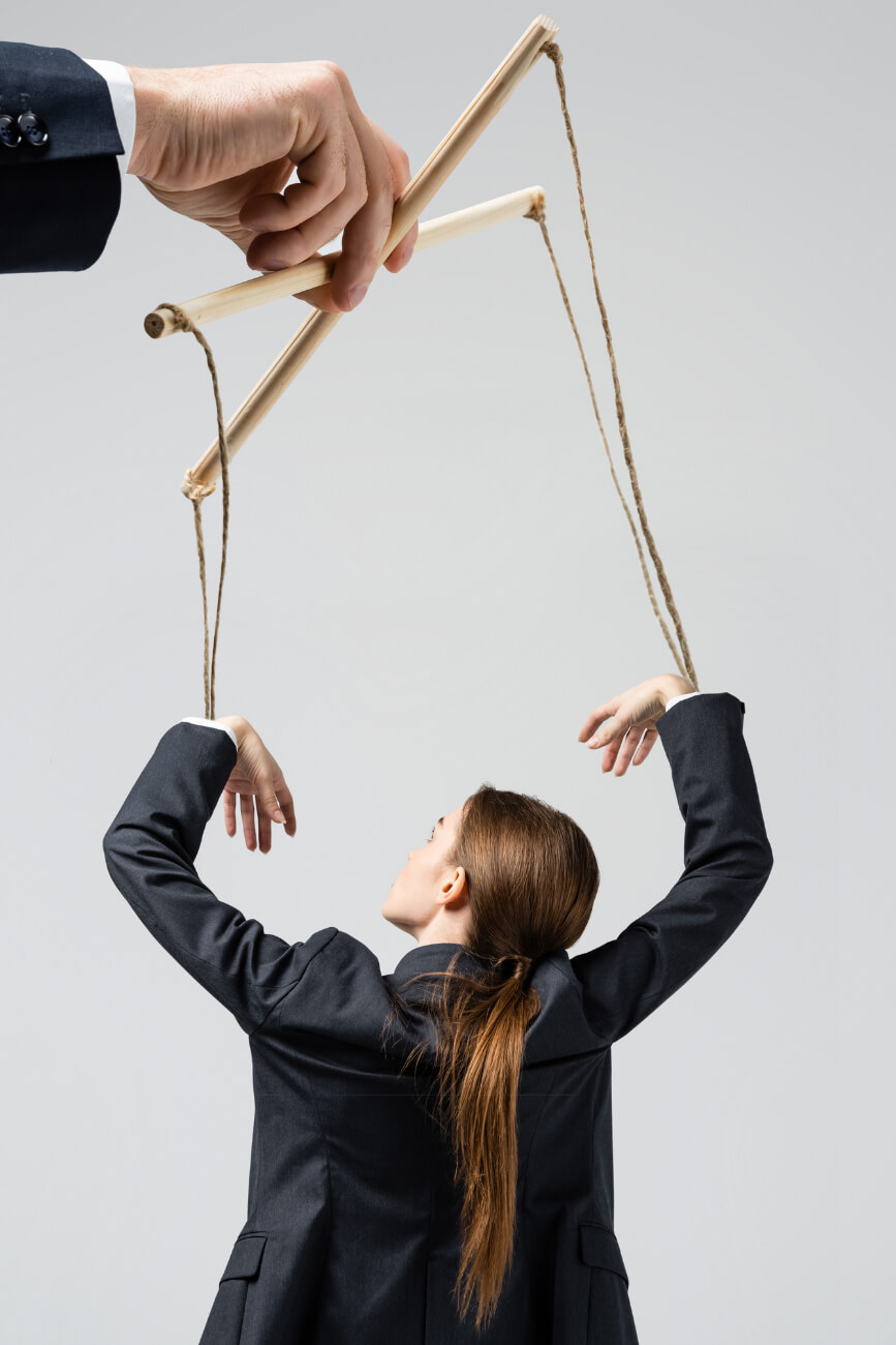 KL Tarot. A woman in a suit is manipulated like a puppet by a giant hand holding strings and wooden sticks, against a neutral background.
